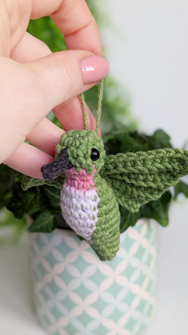 May include: A green, pink and white crocheted hummingbird ornament with black eyes and a black beak. The ornament is hanging from a green string.