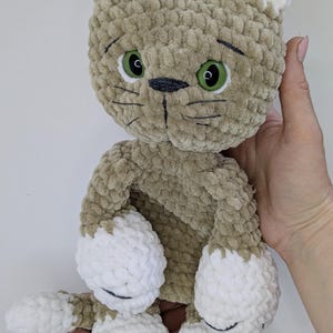 K&ouml;nnte beinhalten: Ein handgemachtes Pl&uuml;schtierspielzeug in Form einer Katze mit hellbraunem K&ouml;rper und wei&szlig;en Akzenten an Ohren, Pfoten und F&uuml;&szlig;en. Die Katze hat gr&uuml;ne Augen und schwarze, gestickte Gesichtsdetails. Das Spielzeug ist aus weichem, strukturiertem Garn gefertigt.