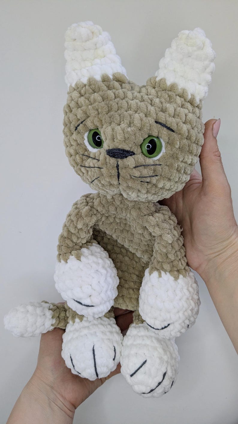 K&ouml;nnte beinhalten: Ein handgemachtes Pl&uuml;schtierspielzeug in Form einer Katze mit hellbraunem K&ouml;rper und wei&szlig;en Akzenten an Ohren, Pfoten und F&uuml;&szlig;en. Die Katze hat gr&uuml;ne Augen und schwarze, gestickte Gesichtsdetails. Das Spielzeug ist aus weichem, strukturiertem Garn gefertigt.