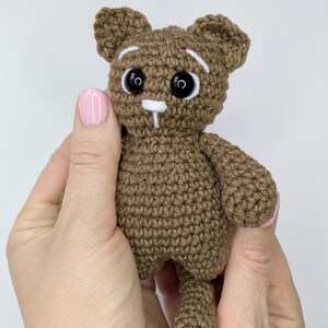 May include: A small brown crocheted cat toy featuring a round head with pointed ears, black plastic safety eyes, white embroidered eyebrows, and a white nose and mouth detail. The body is compact with short arms and a visible tail extending downward.