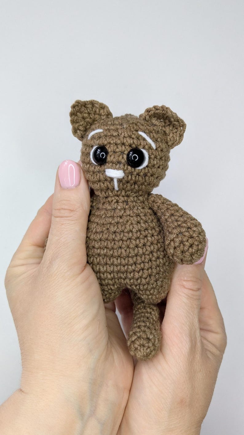 May include: A small brown crocheted cat toy featuring a round head with pointed ears, black plastic safety eyes, white embroidered eyebrows, and a white nose and mouth detail. The body is compact with short arms and a visible tail extending downward.