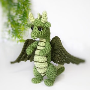 May include: A green crocheted dragon with wings, standing on a white surface. The dragon has a light green belly and dark green wings.
