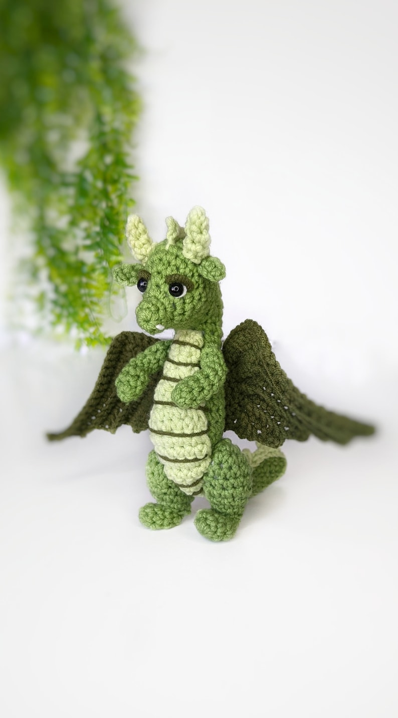 May include: A green crocheted dragon with wings, standing on a white surface. The dragon has a light green belly and dark green wings.