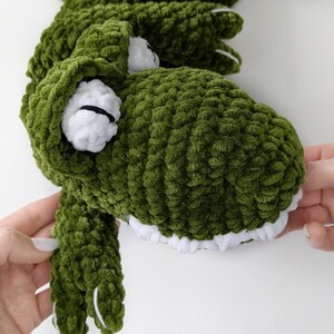 Puede incluir: Un juguete de cocodrilo verde tejido a mano con detalles blancos. El cocodrilo tiene dientes blancos, ojos blancos con pupilas negras y garras blancas. El peluche est&aacute; hecho de un hilo suave y texturizado.