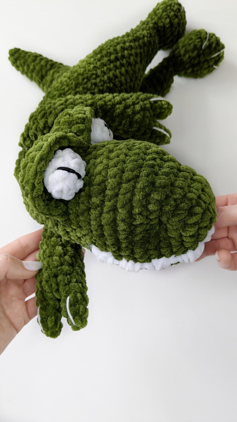 Puede incluir: Un juguete de cocodrilo verde tejido a mano con detalles blancos. El cocodrilo tiene dientes blancos, ojos blancos con pupilas negras y garras blancas. El peluche est&aacute; hecho de un hilo suave y texturizado.