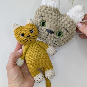 K&ouml;nnte beinhalten: Zwei handgefertigte H&auml;keltierspielzeuge. Eines ist eine senfgelbe Katze mit wei&szlig;en Pfoten und Ohren. Das andere ist ein hellbrauner Katzenkopf mit wei&szlig;en Ohren und gr&uuml;nen Augen. Beide haben schwarze gestickte Details.