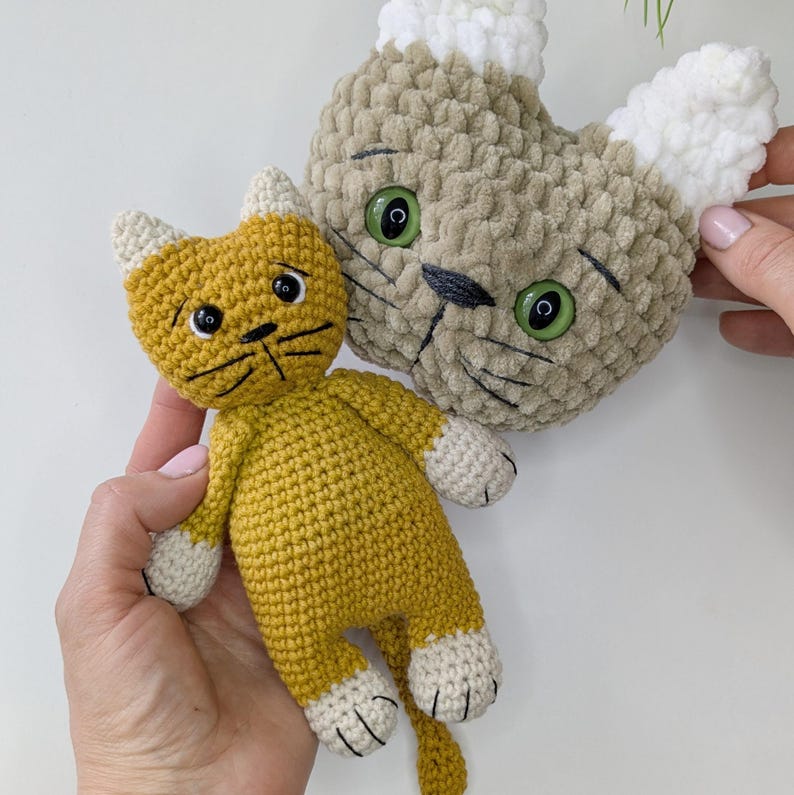 K&ouml;nnte beinhalten: Zwei handgefertigte H&auml;keltierspielzeuge. Eines ist eine senfgelbe Katze mit wei&szlig;en Pfoten und Ohren. Das andere ist ein hellbrauner Katzenkopf mit wei&szlig;en Ohren und gr&uuml;nen Augen. Beide haben schwarze gestickte Details.