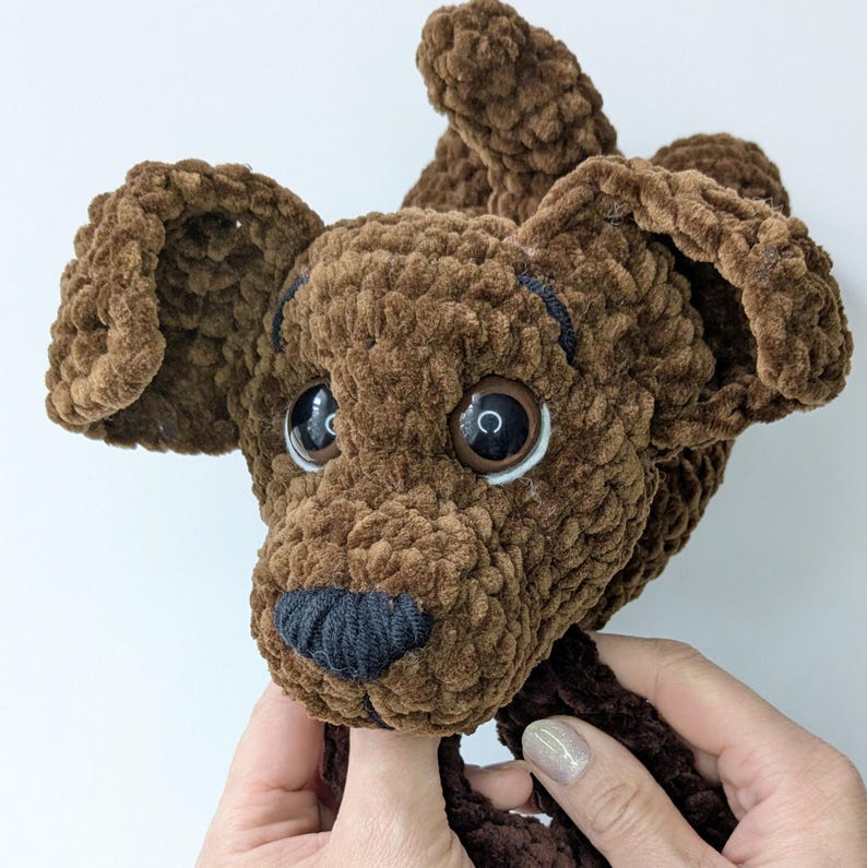 K&ouml;nnte beinhalten: Ein braunes, geh&auml;keltes Pl&uuml;schhund-Spielzeug mit strukturierter Garnoberfl&auml;che, gro&szlig;en dunkelbraunen Kunststoffaugen mit wei&szlig;en Reflexen und einer gestickten schwarzen Nase. Das Stofftier hat Schlappohren und einen runden Kopf.