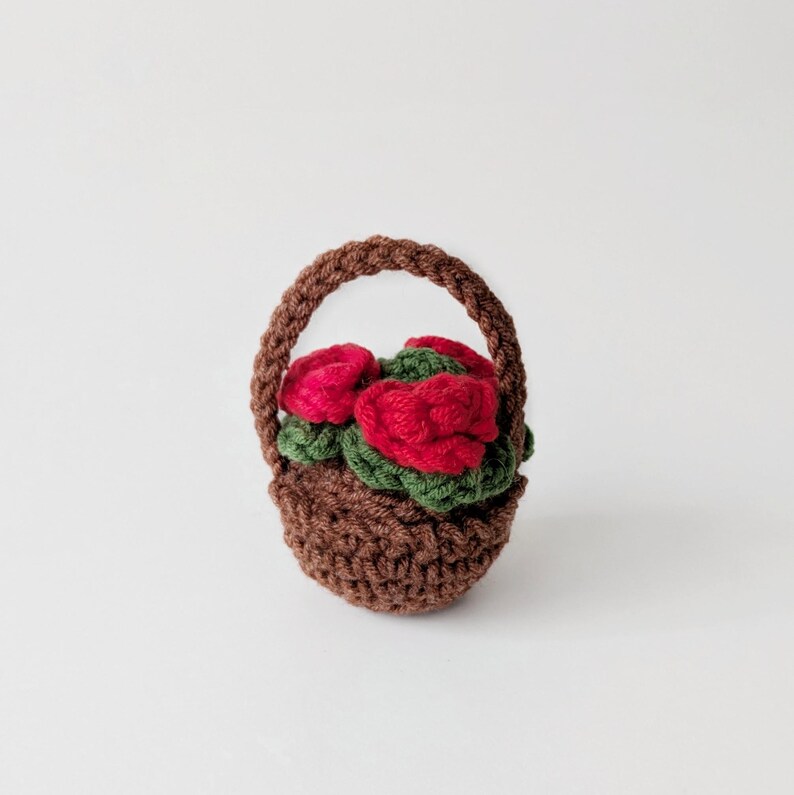 May include: A small, brown crocheted basket filled with red roses and green leaves. The basket has a handle and is made of a textured yarn. The roses are a vibrant red, and the leaves are a deep green.