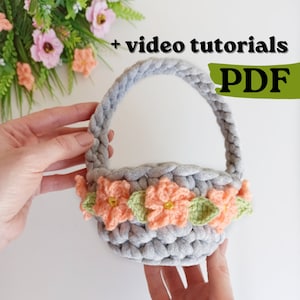 May include: A gray crocheted basket with a handle and a border of peach and green crocheted flowers. The text "video tutorials PDF" is in the upper right corner of the image.