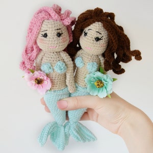 May include: Two crocheted mermaid dolls with light blue tails and beige bodies. The doll on the left has pink hair and the doll on the right has brown hair. Both dolls are wearing light blue seashell bras and holding a flower.