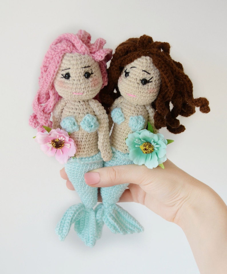 May include: Two crocheted mermaid dolls with light blue tails and beige bodies. The doll on the left has pink hair and the doll on the right has brown hair. Both dolls are wearing light blue seashell bras and holding a flower.