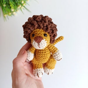May include: A crocheted lion toy with a brown mane, yellow body, and white paws. The lion is being held in a hand.