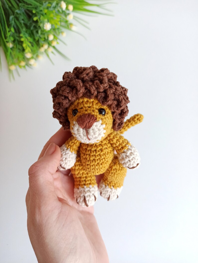 May include: A crocheted lion toy with a brown mane, yellow body, and white paws. The lion is being held in a hand.