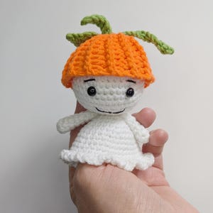 May include: A small crocheted doll featuring a white rounded head and body with a flared base. The doll wears a bright orange textured hat topped with two green crocheted leaves. It has two black bead eyes and a stitched black line for a mouth.