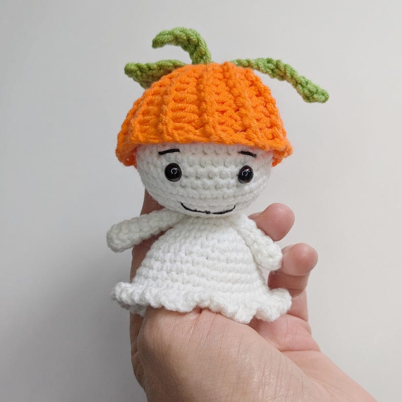 May include: A small crocheted doll featuring a white rounded head and body with a flared base. The doll wears a bright orange textured hat topped with two green crocheted leaves. It has two black bead eyes and a stitched black line for a mouth.