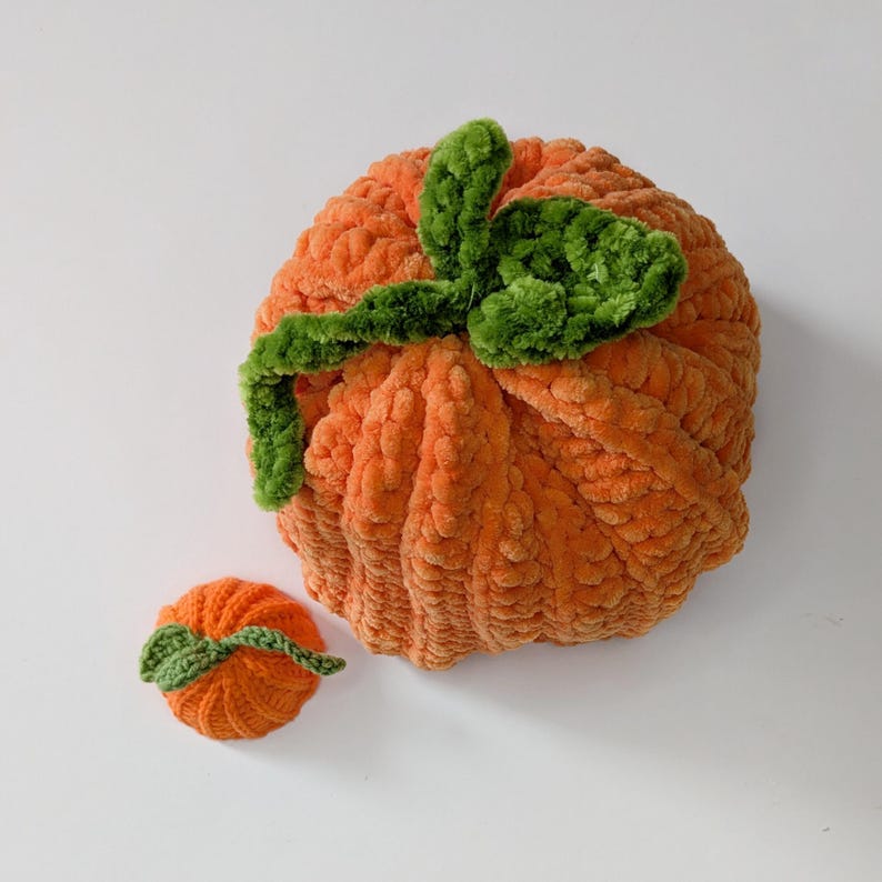 May include: A large and a small orange pumpkin made of soft, textured yarn. Each pumpkin features a ribbed, rounded body and a green crocheted stem with attached green leaves on top, crafted from plush material with a distinct, fuzzy appearance.
