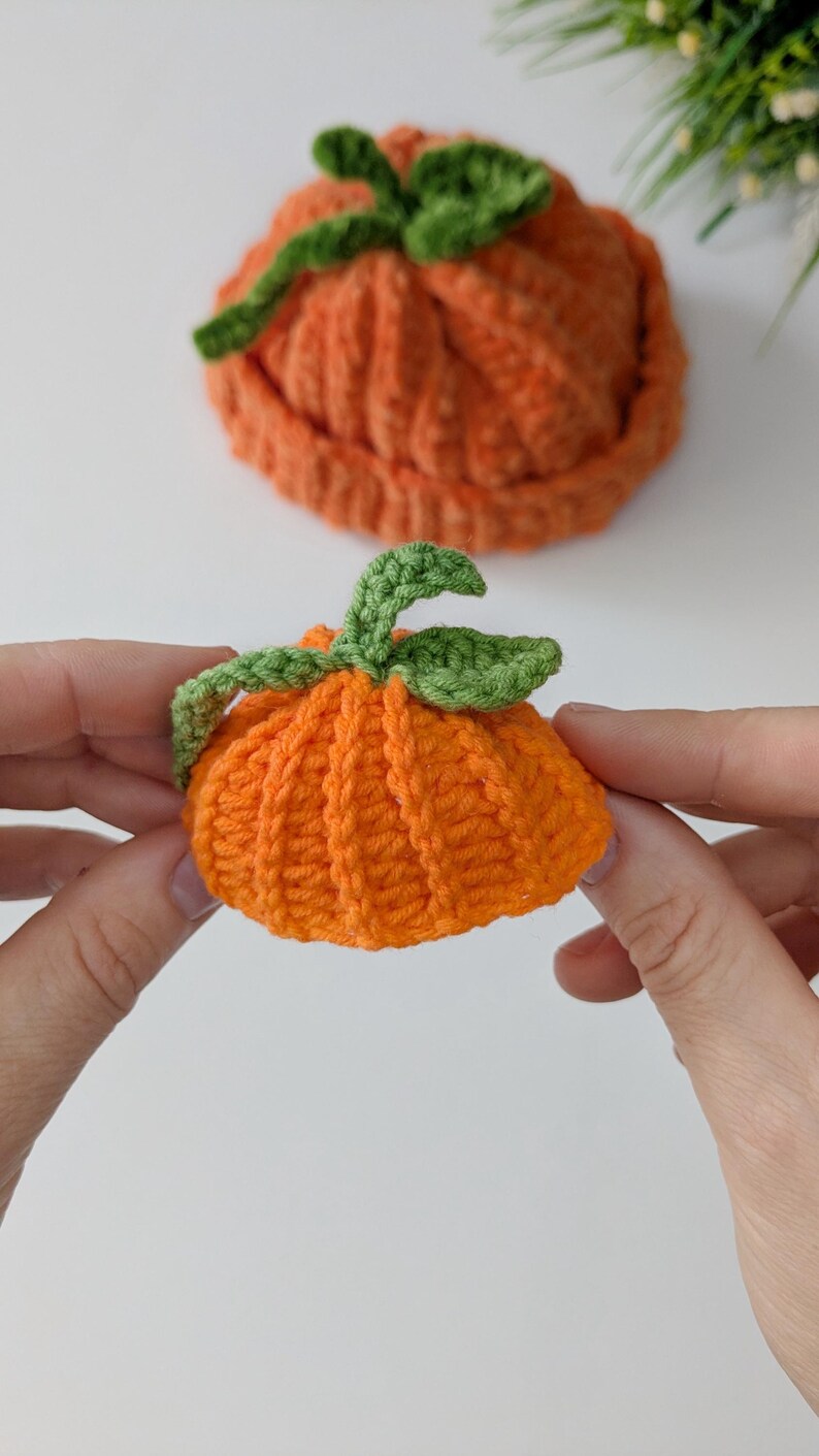 May include: Two small, orange, crocheted pumpkin-shaped items with green crocheted stems and leaves. The items feature a ribbed texture created by the crochet stitch pattern. One pumpkin is held in the foreground, while the other sits behind it.