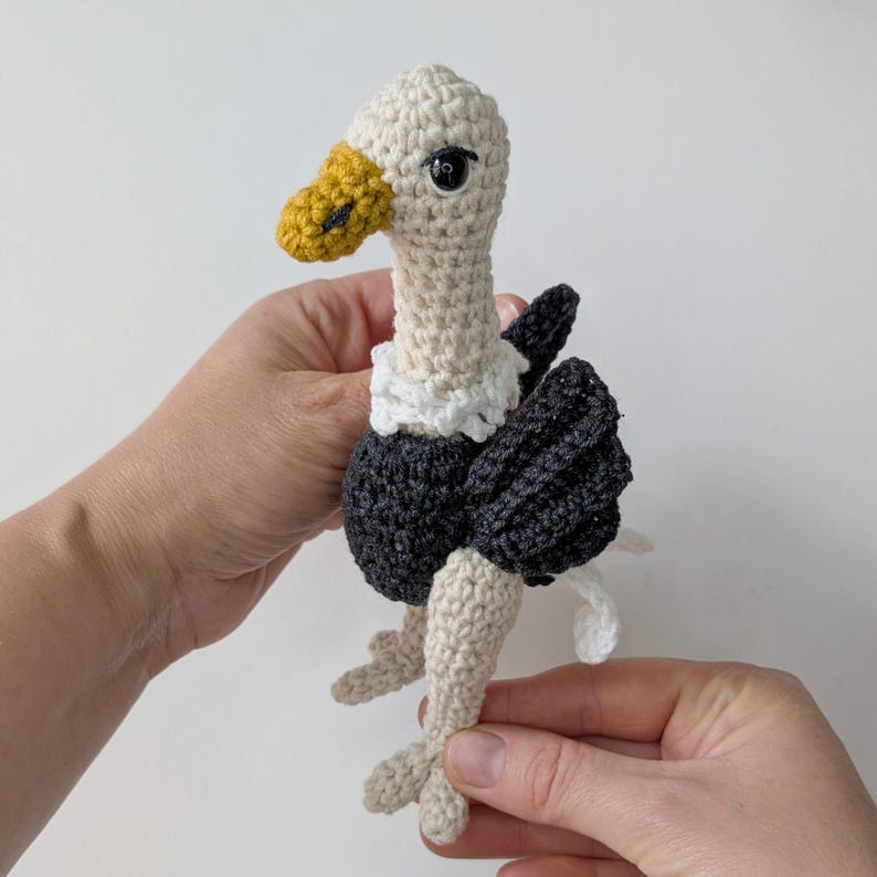 May include: A crocheted vulture toy featuring a cream-colored head and neck, a yellow beak, a white ruff around the neck, and a dark gray body with matching wings. The bird has black plastic eyes and cream-colored legs with three-toed feet.