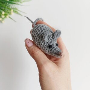May include: A gray crocheted mouse toy with black eyes. The mouse is being held in a hand.