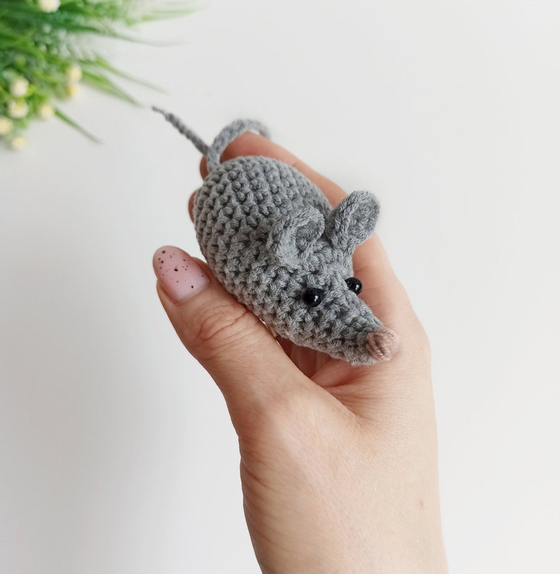 May include: A gray crocheted mouse toy with black eyes. The mouse is being held in a hand.
