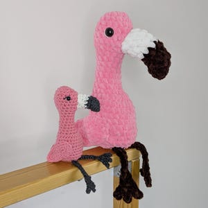 May include: Two handmade crochet flamingo plush toys in different sizes. The larger flamingo is bright pink with a white and dark brown beak and black eyes. The smaller flamingo is a lighter pink with a white and dark gray beak and black eyes.