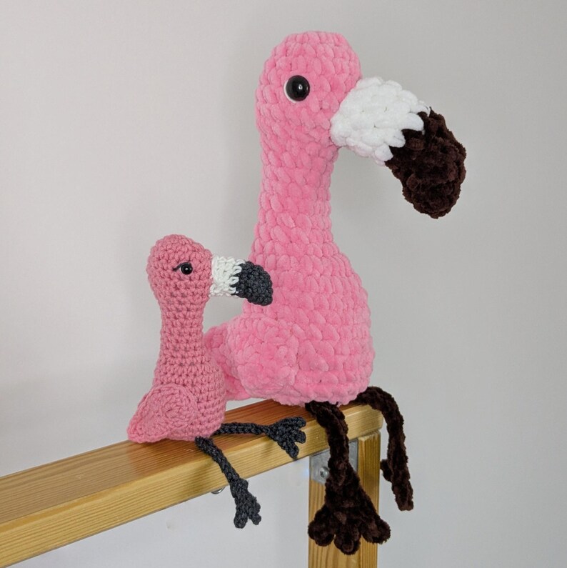 May include: Two handmade crochet flamingo plush toys in different sizes. The larger flamingo is bright pink with a white and dark brown beak and black eyes. The smaller flamingo is a lighter pink with a white and dark gray beak and black eyes.