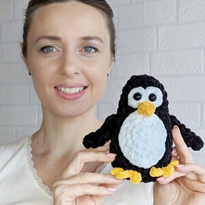 Pode incluir: Uma mulher segura um brinquedo de pinguim de croch&ecirc; preto e branco com p&eacute;s amarelos. O pinguim tem olhos pretos grandes e um bico laranja brilhante.