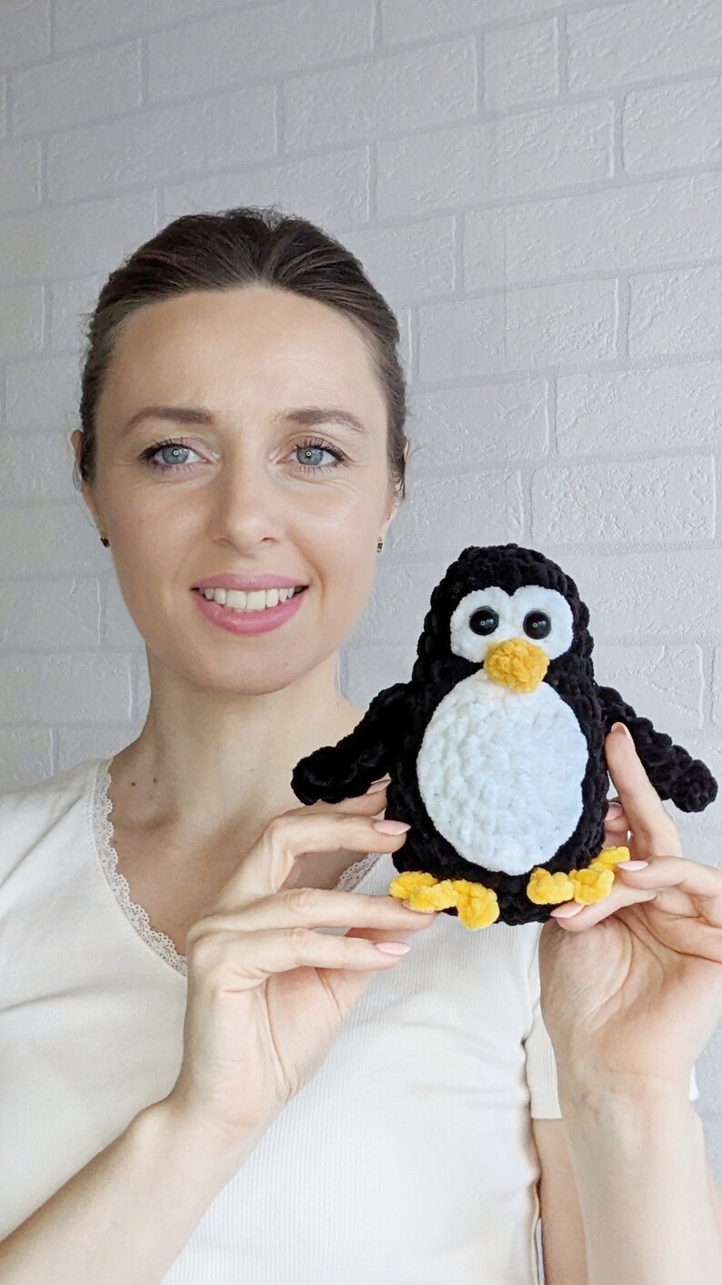 Pode incluir: Uma mulher segura um brinquedo de pinguim de croch&ecirc; preto e branco com p&eacute;s amarelos. O pinguim tem olhos pretos grandes e um bico laranja brilhante.