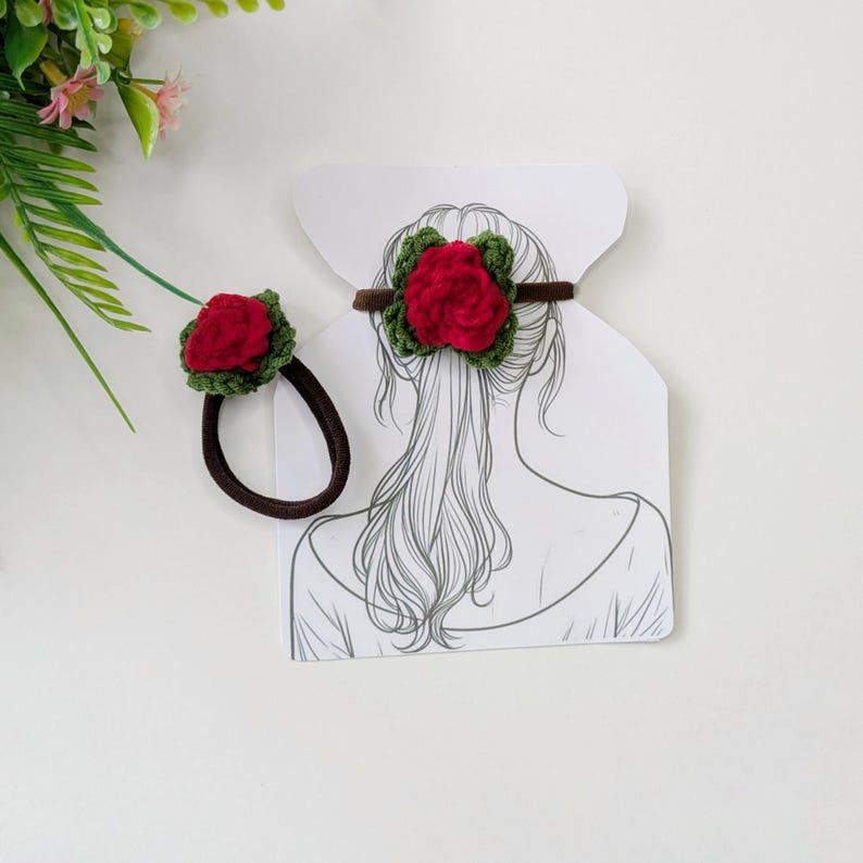 May include: Two handmade hair accessories: a red rose crochet hair tie and a matching elastic hair band. The red roses are surrounded by green leaves. The hair tie is brown and the elastic band is black. The accessories are displayed on a white background.