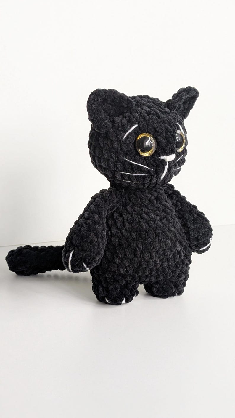 May include: A black crocheted cat plush toy made from soft, textured yarn. The figure features large, round, amber-colored eyes with a glittery finish, white embroidered whiskers, a small white nose, and white stitched details on the paws.