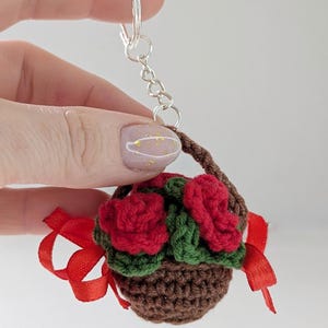 May include: A crocheted keychain featuring a miniature basket filled with red roses and green leaves. The brown basket has a handle and is adorned with a red ribbon. The keychain is attached to a silver ring.