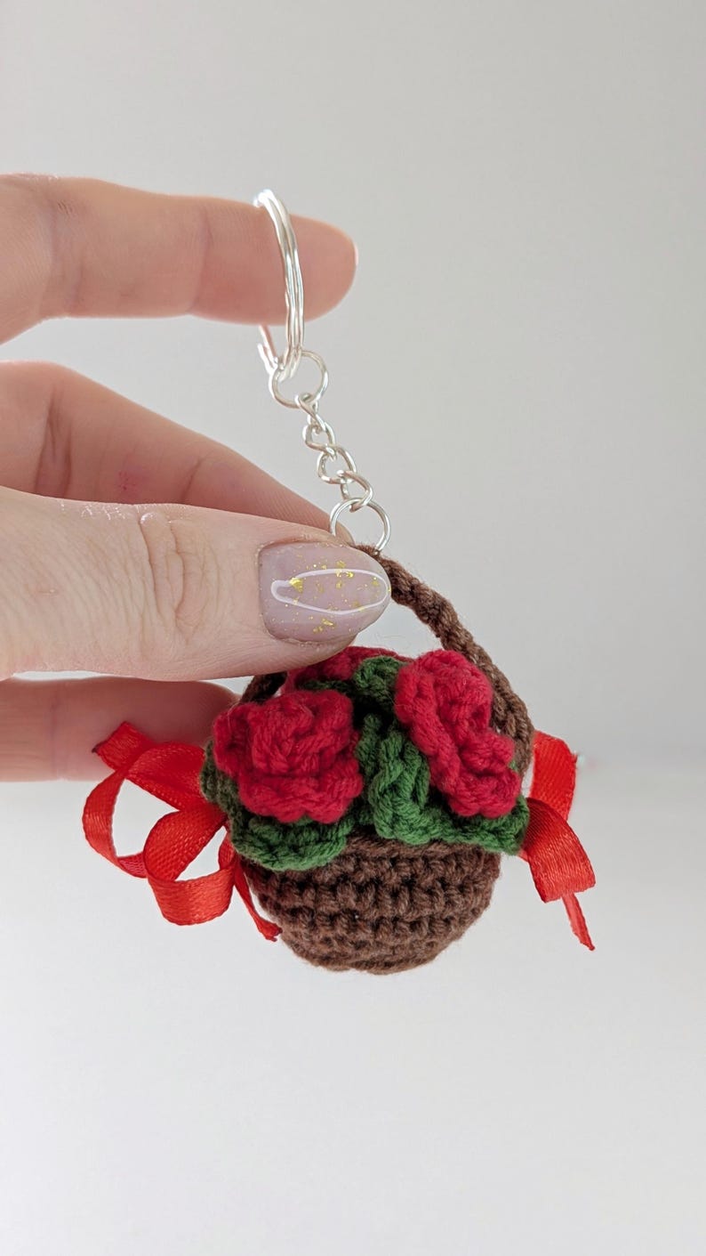 May include: A crocheted keychain featuring a miniature basket filled with red roses and green leaves. The brown basket has a handle and is adorned with a red ribbon. The keychain is attached to a silver ring.