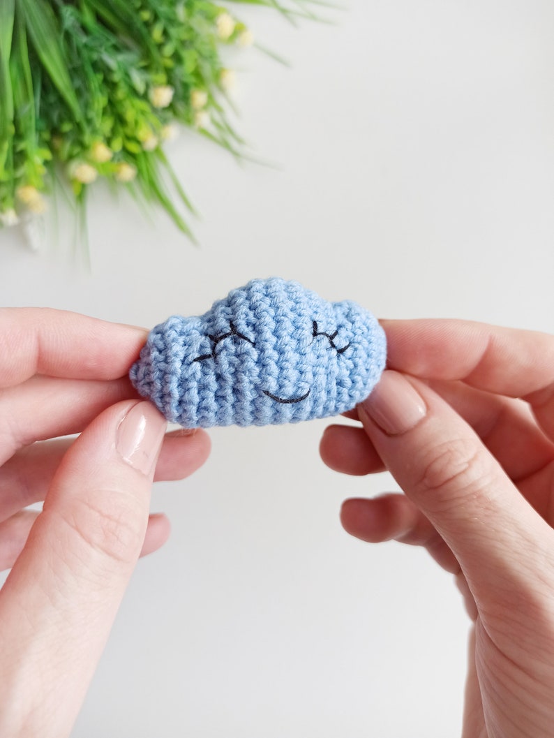 May include: A light blue crocheted cloud with a smiling face. The cloud is being held in two hands.