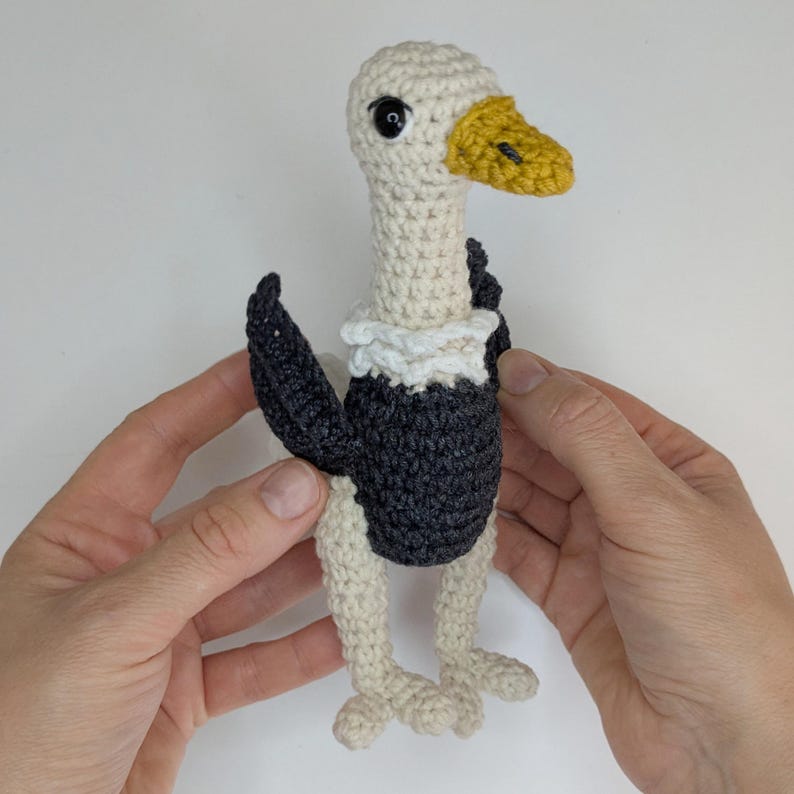 May include: A crocheted vulture toy featuring a cream-colored head and neck, a yellow beak, a white ruff around the neck, and a dark gray body with matching wings. The bird has black plastic eyes and cream-colored legs with small feet.