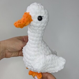May include: A handmade crochet plush duck made from soft white yarn with a textured, fuzzy appearance. The duck features a bright orange beak and matching orange feet, along with a single black plastic safety eye visible on the side of its head.