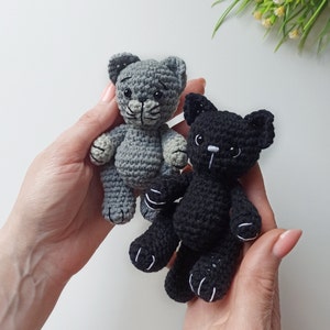 May include: Two crocheted cat toys, one gray and one black, with white accents on the eyes and paws. The toys are being held in the hands of a person.
