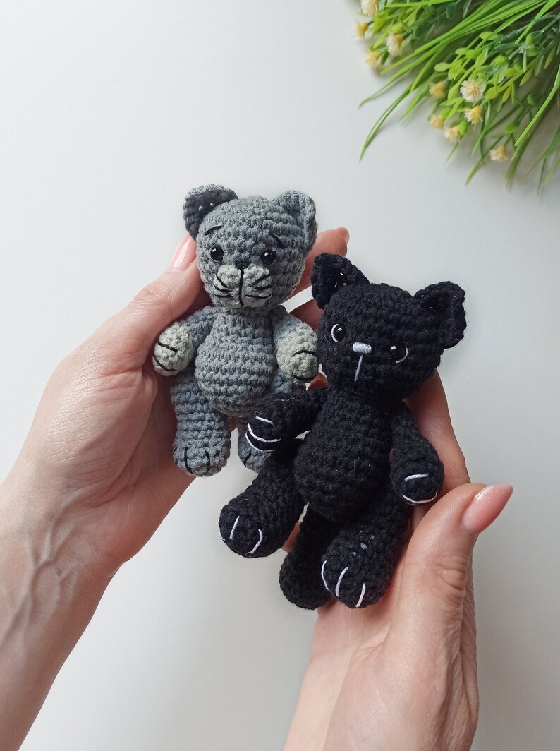 May include: Two crocheted cat toys, one gray and one black, with white accents on the eyes and paws. The toys are being held in the hands of a person.