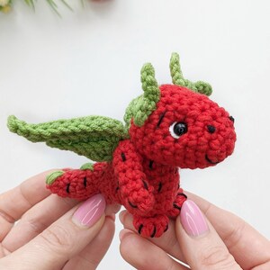 May include: A crocheted strawberry dragon toy with green leaves for wings and a black eye. The dragon is held in two hands.