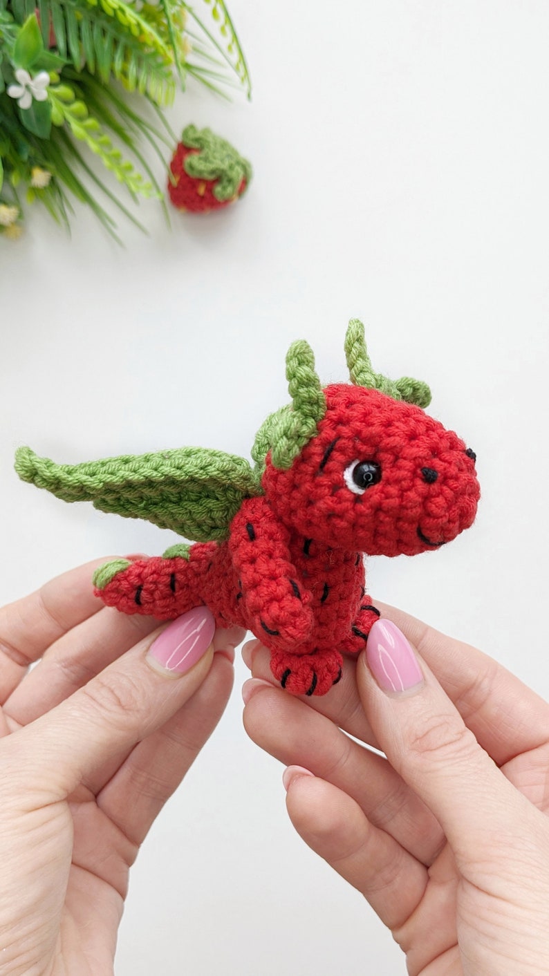 May include: A crocheted strawberry dragon toy with green leaves for wings and a black eye. The dragon is held in two hands.