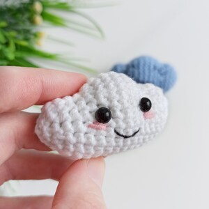 May include: A white crocheted cloud with a smiling face, black eyes, and pink cheeks. The cloud is being held in a hand. A blue crocheted cloud is visible in the background.