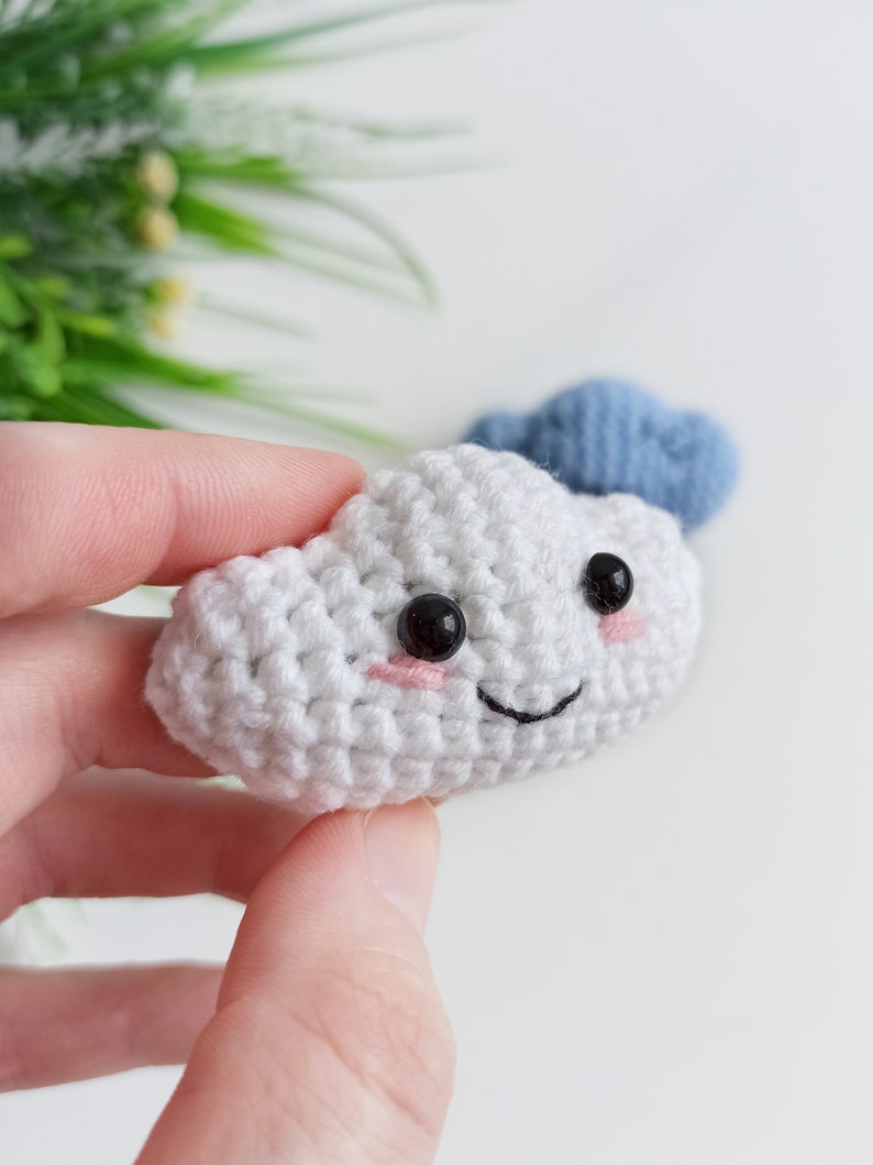 May include: A white crocheted cloud with a smiling face, black eyes, and pink cheeks. The cloud is being held in a hand. A blue crocheted cloud is visible in the background.