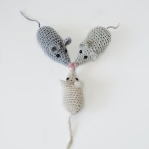 May include: Three crocheted mice toys, two gray and one beige, with black eyes and pink noses. The mice are arranged in a triangle shape.