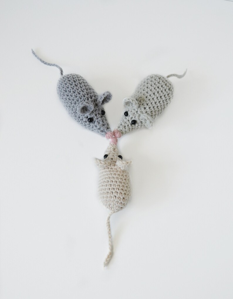 May include: Three crocheted mice toys, two gray and one beige, with black eyes and pink noses. The mice are arranged in a triangle shape.