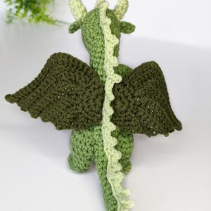 May include: A green crocheted dragon with light green accents. The dragon has wings and a long tail.