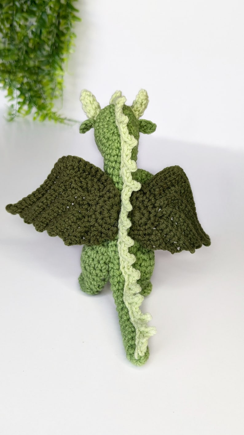 May include: A green crocheted dragon with light green accents. The dragon has wings and a long tail.