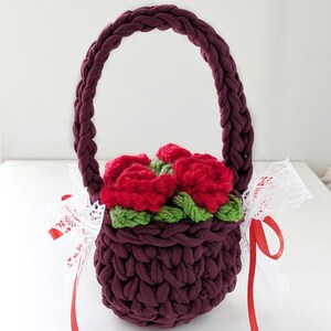 May include: A burgundy crochet basket with a braided handle, filled with red crochet roses and green leaves. White lace and red ribbon are tied to the basket. The basket is a handmade item.