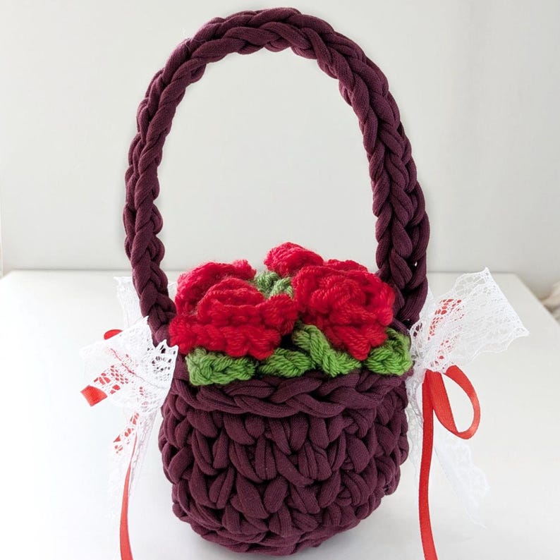 May include: A burgundy crochet basket with a braided handle, filled with red crochet roses and green leaves. White lace and red ribbon are tied to the basket. The basket is a handmade item.