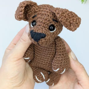 K&ouml;nnte beinhalten: Ein kleines geh&auml;keltes Hundespielzeug in Braun mit gro&szlig;en schwarzen Kunststoffaugen, einer schwarzen gestickten Nase und Schlappohren. Die Garnstruktur ist am ganzen K&ouml;rper sichtbar, mit kleinen wei&szlig;en Stickdetails an den Pfoten und dunklen Augenbrauen.