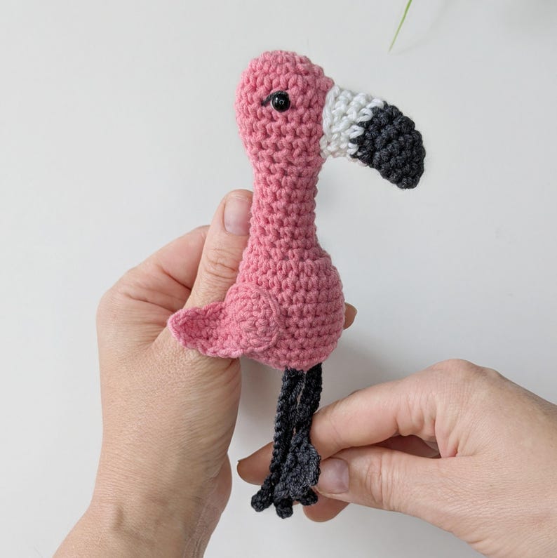 May include: A small, handmade crochet flamingo toy featuring a bright pink body, a white and dark gray curved beak, and dark gray legs. The flamingo has a single black bead eye on the side of its head and a small, textured wing on its side.